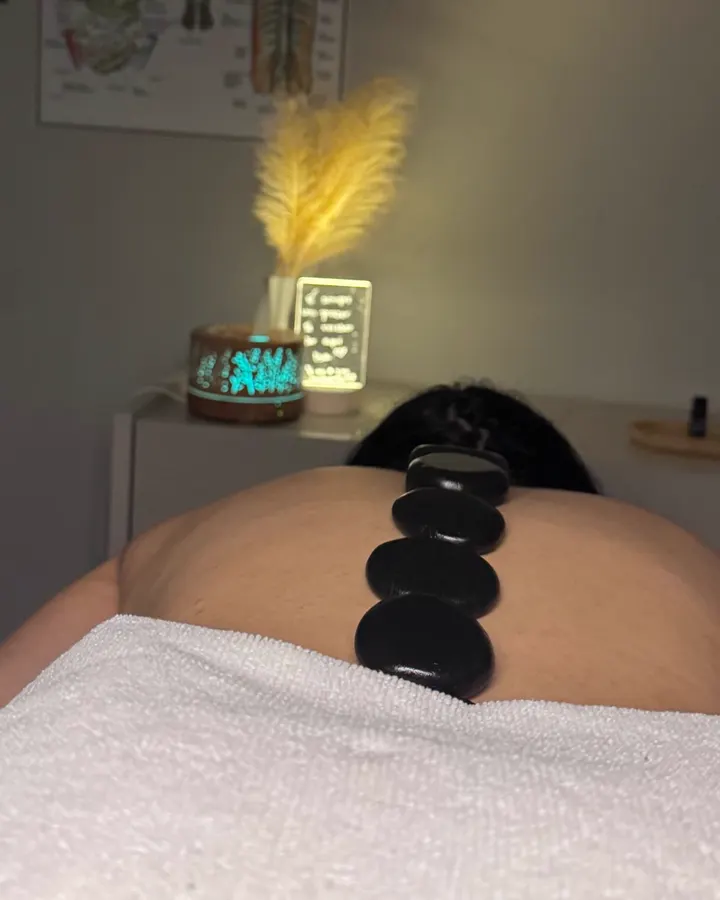 Pedras quentes alinhadas nas costas de uma cliente, com luz de abajur e pluma de pampas ao fundo